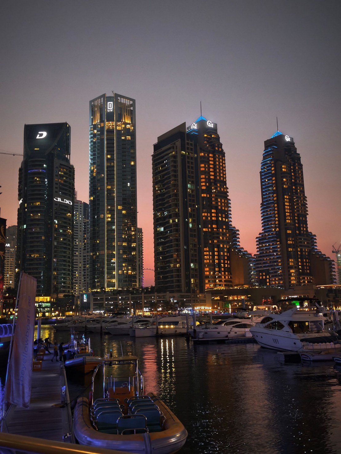 Dubai Marina towers at dusk, with boats moored along the waterfront.