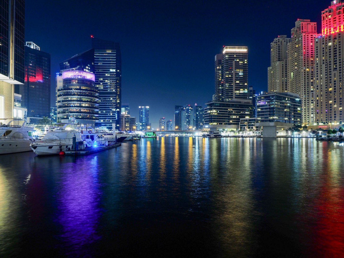Dubai Marina at night: residential towers lit up and reflected in the water, with yachts moored along the left bank.